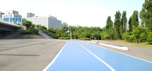 blue running track, on a city park, in Seoul, a paved area with a blue-painted section, surrounded by lush green trees and bushes. buildings and structures, on a sunny day with a clear sky