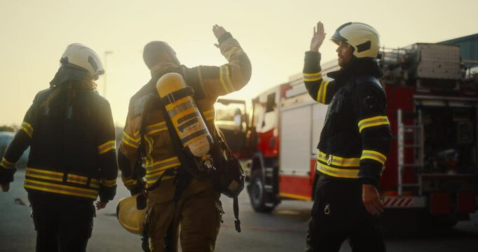 Portrait of Three Diverse Firefighters Dressed in Their Full Gear Giving Each other a High Five Outside an Emergency Location After the Fire is Secured. Fire Truck Parked Next to Them on a Street
