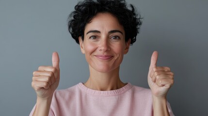 A cheerful woman dressed in a pink top stands indoors, giving a thumbs-up with both hands, radiating positivity and satisfaction against a neutral background.