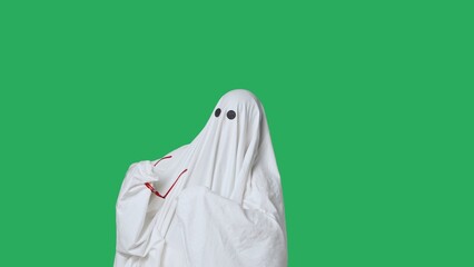 Funny halloween ghost in white sheet takes off red glasses looks around, isolated on chroma key green screen background. Happy halloween spooky concept.