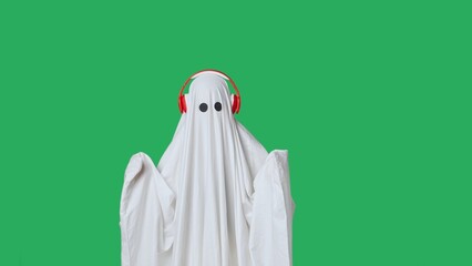 Funny halloween ghost in white sheet wearing headphone dancing listening music, isolated on chroma key green screen background. Happy halloween spooky concept