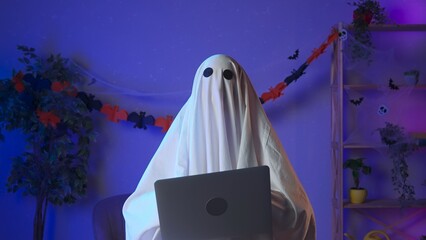 Funny halloween ghost in white sheet with black eyes in dark room working on laptop, typing text...