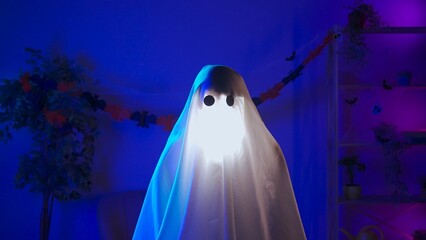 Funny halloween ghost in white sheet with black eyes in dark room and lighted up a flashlight under...