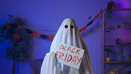 Funny halloween ghost in white sheet with black eyes in dark room holding showing paper sign with...