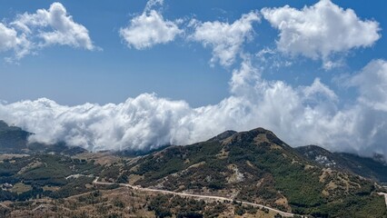 A sweeping view of a mountainous landscape with lush green hills, partially covered by low-hanging clouds, set against a bright blue sky with scattered clouds.