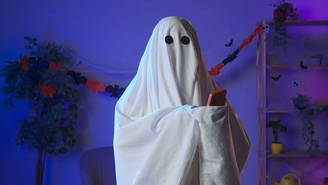 Funny halloween ghost in white sheet with black eyes in dark room talking on smartphone by video call, positive expression. Happy halloween spooky concept.
