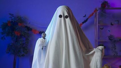 Funny halloween ghost in white sheet with black eyes in dark room dancing to music and sings in karaoke microphone. Happy halloween spooky concept.