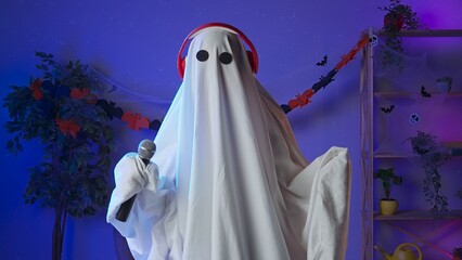 Funny halloween ghost in white sheet with black eyes in dark room dancing to music in headphones...