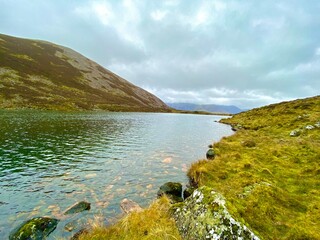 A tranquil mountain lake bordered by grassy slopes and rugged hills under an overcast sky, reflecting the untouched beauty of nature.