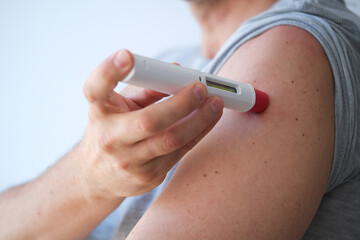 an anonymous adult man injecting himself a treatment such as insulin or Adalimumab, Etanercept or epinephrine into his shoulder. It is a white pen injector with red head for health care