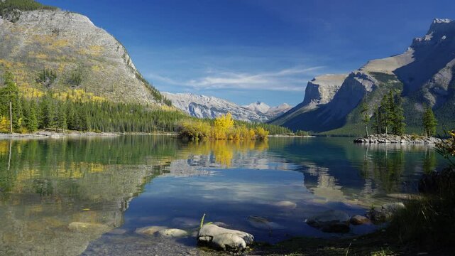 Banff National Park Lake Minnewanka autumn landscape. Alberta, Canada. Majestic mountains, forests in yellow green color against blue sky reflected on the water. Beautiful natural scenery.