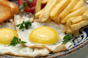 Breakfast plate with fried eggs, sausage, tomatoes, and fries served on a decorative blue plate