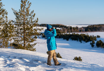 woman birdwatcher in a blue jacket scans the snowy winter landscape and forest with binoculars,...