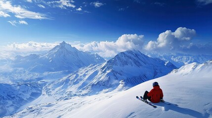 A skier resting at the top of a snowy slope, looking out over snow-capped mountains and a deep blue winter sky.