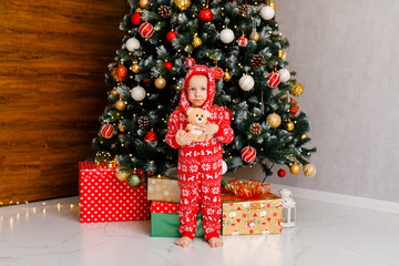 2-year-old little boy in warm pajama is holding teddy bear next to Christmas tree with gifts