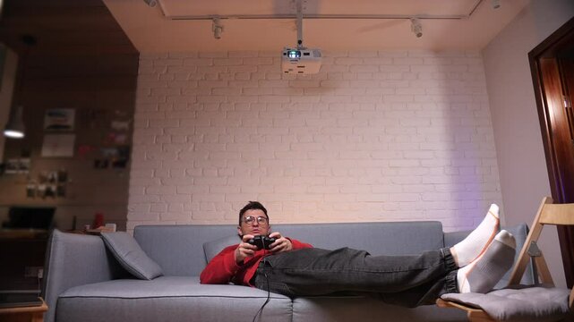 gamer man lying on sofa and playing video games on console using controller