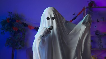Funny halloween ghost in white sheet with black eyes in dark room dancing to music and sings in karaoke microphone. Happy halloween spooky concept.