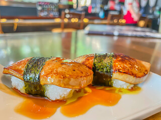 Close-up of two nigiri sushi, topping with foie gras. The elegant presentation is enhanced by the light reflections from the background, suggesting a vibrant sushi bar atmosphere.