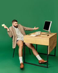 Young man in business suit on top sitting at desk in underwear with his yellow credit card and spreading hands against green studio background. Concept of shopping, sales, Black Friday, Cyber Monday