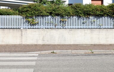 Fence made of concrete wall with metal railing with large slats on top. Building and hedge on...