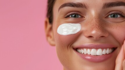 Fototapeta premium A smiling woman applying facial cream, showcasing skincare routine on a pink background, promoting beauty and self-care.
