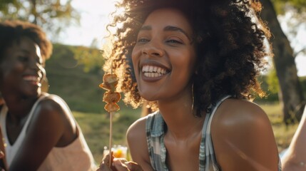 Fototapeta premium A woman enjoying an outdoor picnic on a sunny day, holding a skewer with grilled food, laughing heartily with friends against a backdrop of greenery.