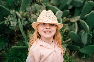 Red-haired happy toothless seven-year-old girl in hat enjoys life and smiles standing against cacti...