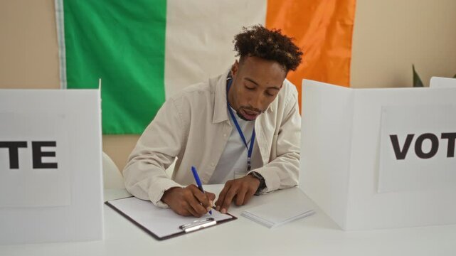 A young black man sits indoors in an electoral college room with the irish flag behind him, writing on a document between two vote signs. - Powered by Adobe