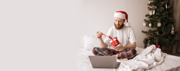Excited young man unpacking Xmas present pulling ribbon when sitting on bed at home, opening gift box with excitement during christmas morning