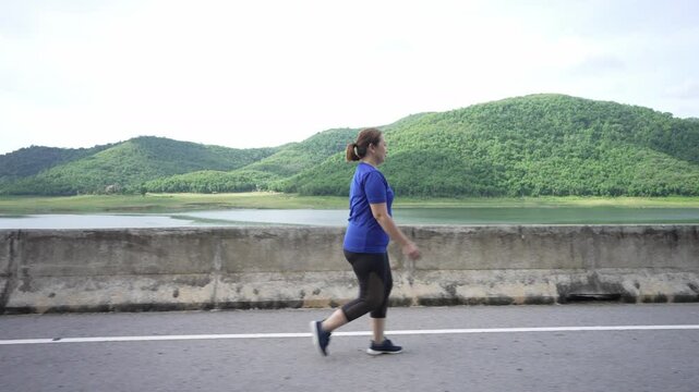 A side view of a middle-aged, overweight Asian woman walking for exercise on a scenic road by a reservoir, aiming for better health and weight loss