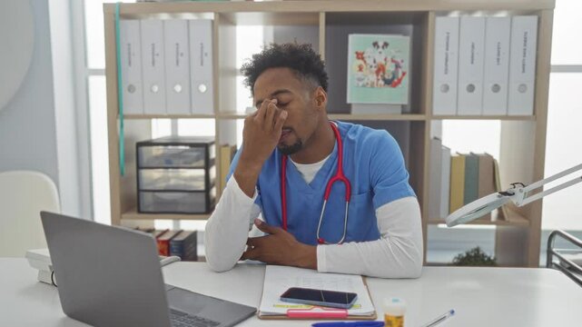 A young, african american man with a beard and stethoscope sits stressed at a desk in a veterinary clinic with a laptop, smartphone, documents, and a colorful animal poster in the background.