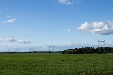 Bright green meadow withrow of high-voltage power line poles in the background against the blue sky with white clouds. Power lines stretching into the distance. Perspective. Electricity