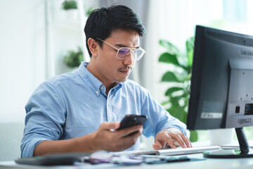 A man wearing glasses is sitting at a desk with a computer monitor and a cell phone. He is looking at the cell phone while typing on the computer