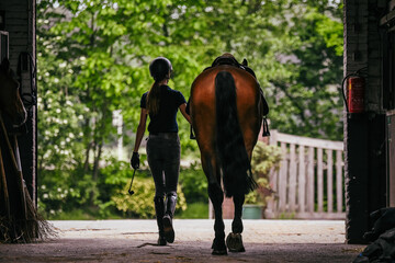 The back of the rider, walking towards the exit with her horse by her side with saddle on, ready to ride