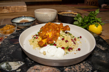 A delicious plate of slow cooked melt in the mouth Chicken Tagine, with couscous surrounded by the ingredients used to make the dish on a rustic wooden kitchen work top