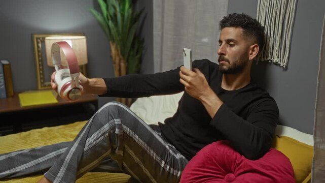 Handsome young hispanic man relaxes in his bedroom, taking a photo of his headphones with his phone while lying on the bed.