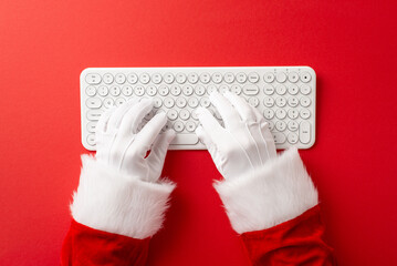 Santa Claus hands in gloves typing on a white keyboard against a red background symbolizing holiday preparations and Christmas communication