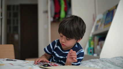 Young boy deeply engaged with a small toy or puzzle on his bed, situated in a child’s room filled with books and toys, highlighting a moment of solitary play and focus