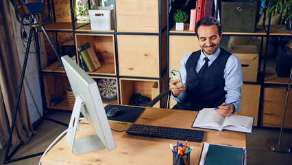 Businessman smiling while using his phone, scrolling through social media instead of focusing on work laid out in front of him. Concept of business, people and daily work challenges. Ad.