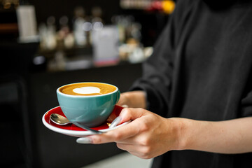 A close-up of hands holding a teal cup and saucer with a cappuccino featuring heart-shaped latte art. The image conveys warmth and hospitality, perfect for illustrating café culture or barista skills.