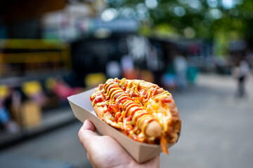 close-up of a hand holding a hot dog with condiments in a paper tray, set against a blurred street food market background. The image captures the vibrant atmosphere and appeal of street food culture.