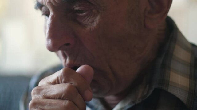Caucasian pensioner 70-79 years old coughs heavily into his fist while inside, portrait of an elderly man coughing