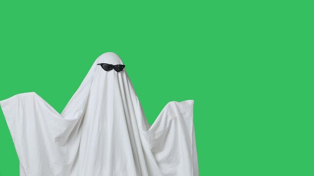 Funny halloween ghost in white sheet wears black glasses dancing coolly, isolated on chroma key green screen background. Happy halloween spooky concept.