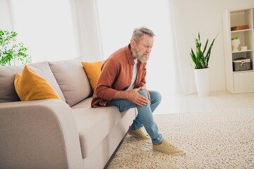 Full length photo of nice pensioner man sit couch touch knee pain dressed brown garment spend pastime spacious house indoors room