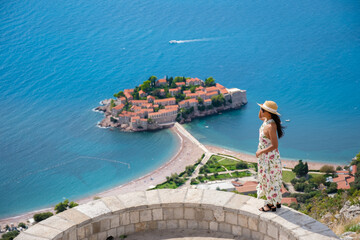 A woman stands on a stone balcony, gazing at the stunning island of Sveti Stefan, surrounded by...
