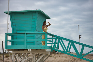 Pretty young woman with green eyes in a yellow silk suit and pink heels, hand over her eyes looking at the sea, leaning on a railing of a green lifeguard hut. Concept beauty, beach, coast, ocean.