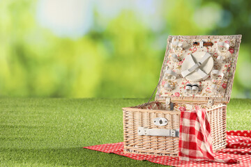 One picnic wicker basket with checkered napkin, tableware and blanket on grass against blurred green background. Space for text