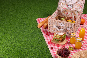 Fototapeta premium Picnic basket with tableware, different snacks, juice and hat on green grass outdoors, space for text