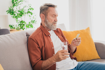 Photo of nice pensioner man sit couch water glass take meds dressed brown garment spend pastime spacious house indoors room