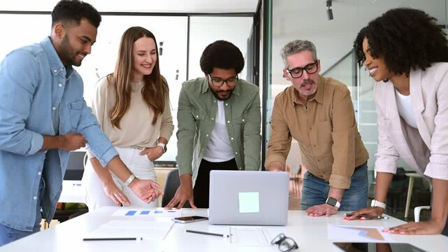 Diverse group of professionals leans in towards a laptop, sharing a moment of collaboration and interaction in their modern workspace. The energy and engagement of a diverse team working on a project